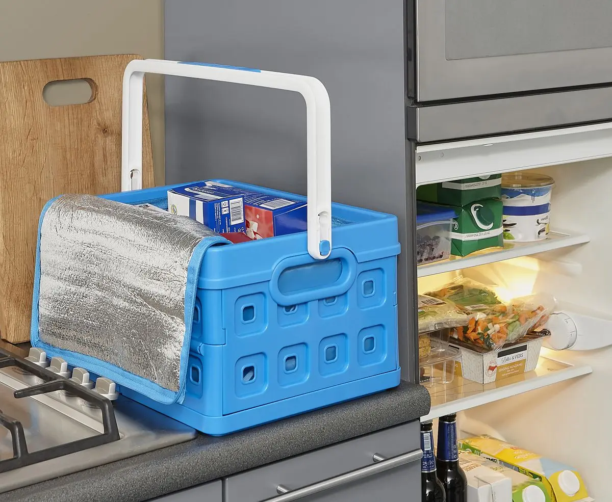 Blaue Faltkiste mit gekühlten Einkäufen steht neben einem geöffnetem Kühlschrank
